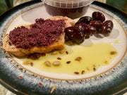 Liquid Gold Products' organic kalamata olive tapenade made with organic oregano, greek extra virgin olive oil and black pepper, on wholemeal bread, with olives, olive oil and seeds, on a plate